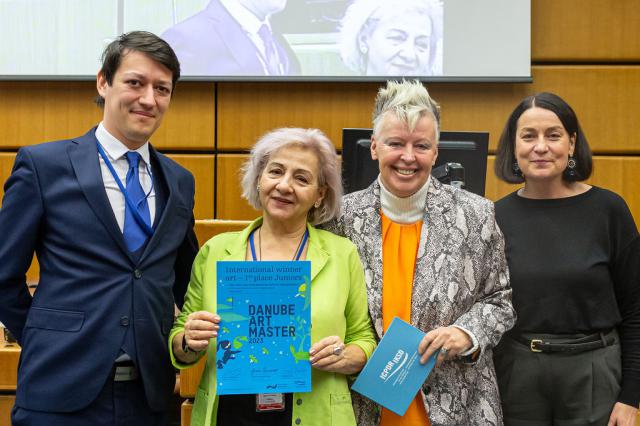 4 persons in a large meeting room in from of a large screen, the second from left a woman holding a blue certificate